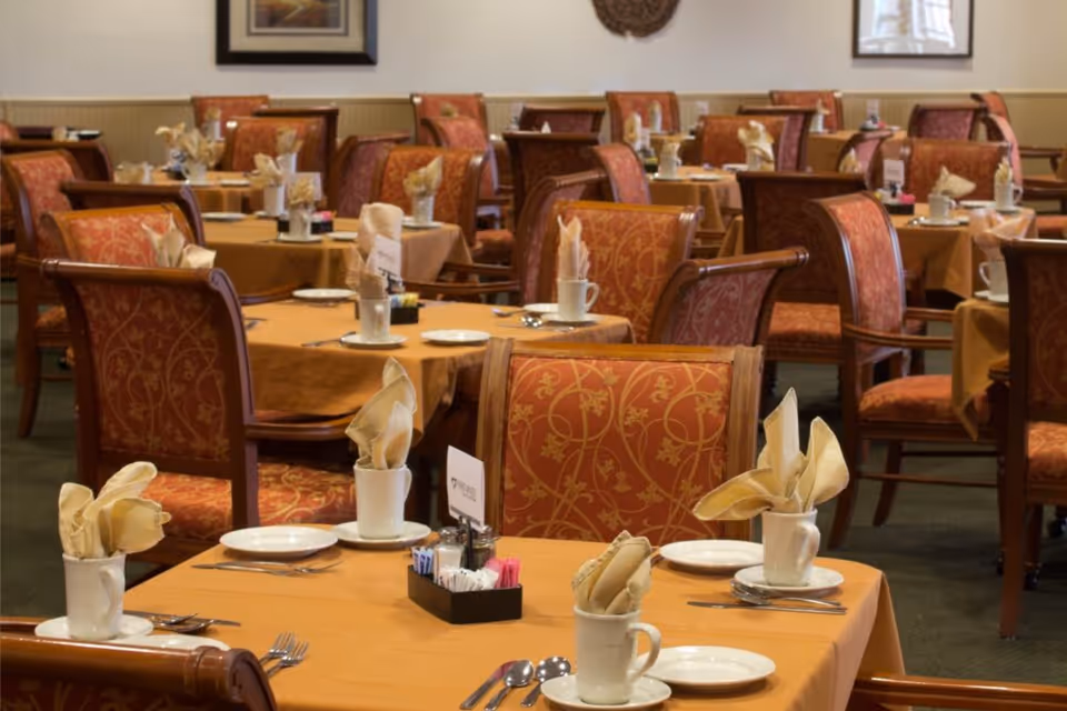 Dining room with multiple set tables covered in orange tablecloths, folded napkins in mugs, and patterned upholstered chairs.