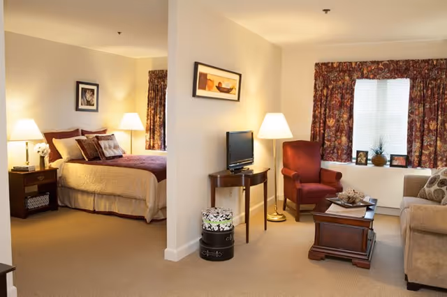 A cozy senior living apartment with a bedroom on the left featuring a bed with pillows and bedside tables with lamps. On the right is a living room area with an armchair, a sofa, a coffee table, a small TV on a wooden table, and a window with floral curtains.