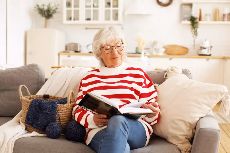 An elderly woman with white hair and glasses wearing a red and white striped sweater sits on a gray couch reading a book. Next to her is a woven basket with blue yarn and knitting needles. The background shows a bright, cozy kitchen with white cabinets and wooden countertops.