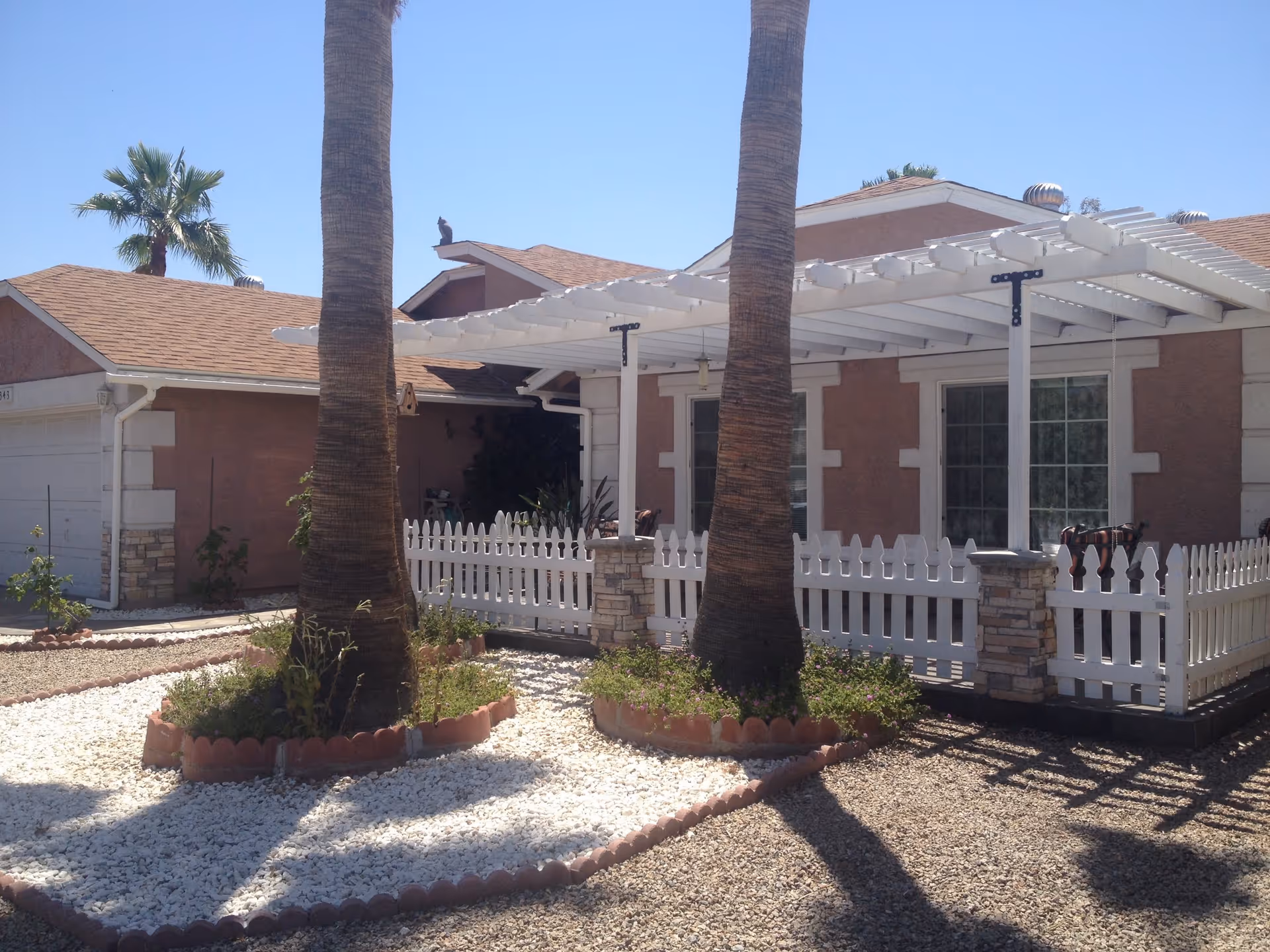 Exterior view of a single-story residential building with a white picket fence and a white pergola over a patio area. Two tall palm trees are planted in small garden beds surrounded by white gravel in the front yard. The building has a brownish exterior with white trim and a garage on the left side. The sky is clear and blue.