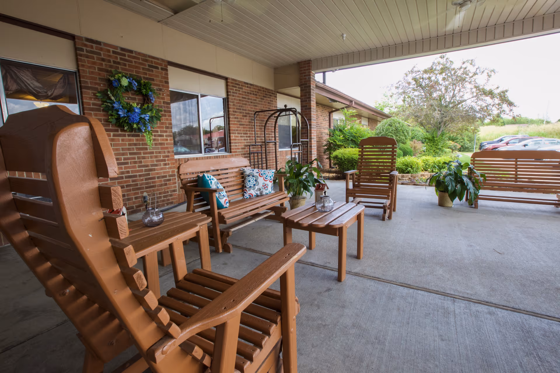 Covered outdoor patio area with wooden rocking chairs, benches, and a small table. The space is decorated with potted plants and a wreath on the brick wall. There is greenery and parked cars visible in the background.
