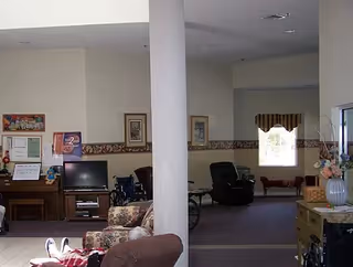 Interior view of a senior living facility common area with various seating options including armchairs and a floral-patterned sofa. There is a white pillar in the center, framed pictures on the walls, a piano with certificates and decorations on top, and a window with a valance in the background. A wheelchair is also visible near the seating area.