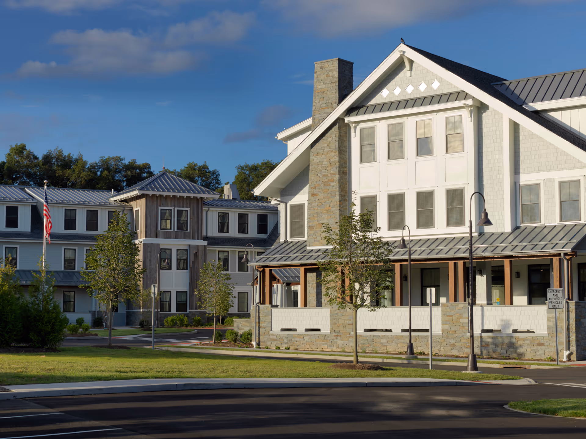 Exterior view of a senior living facility building with a stone chimney, multiple windows, and a covered porch area. The building is surrounded by small trees and a well-maintained lawn under a blue sky with some clouds.