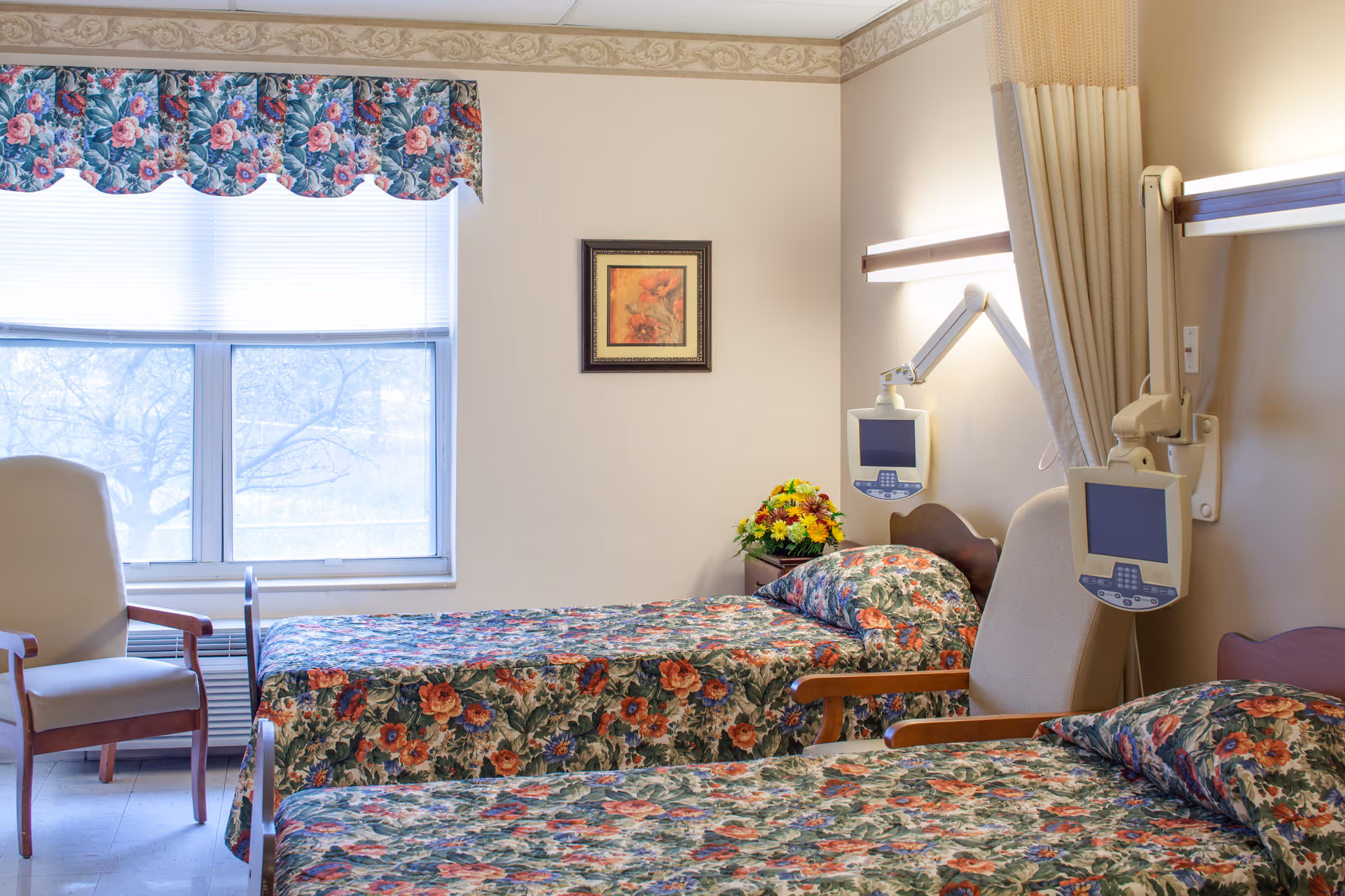 A nursing facility patient room with two twin beds covered in floral bedspreads, chairs, wall-mounted monitors and a window.