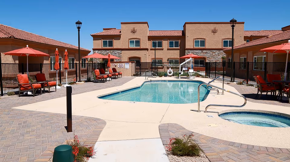 Outdoor swimming pool area at a senior living facility with lounge chairs, red umbrellas, and a hot tub. The pool is surrounded by a paved patio and a black metal fence, with a two-story building in the background under a clear blue sky.