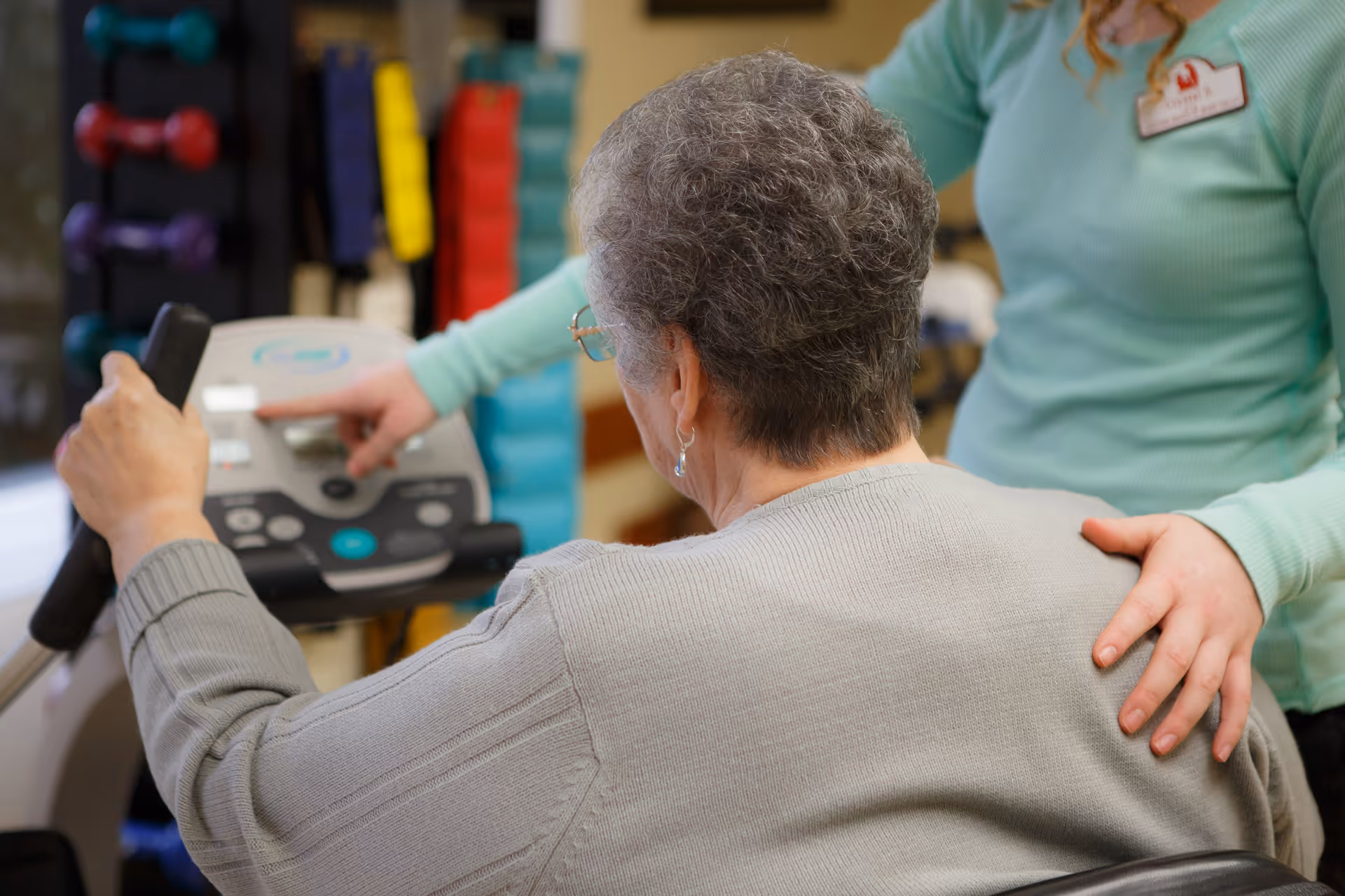An elderly woman using an exercise machine with the assistance of a caregiver who is pointing at the machine's controls and gently supporting her back.