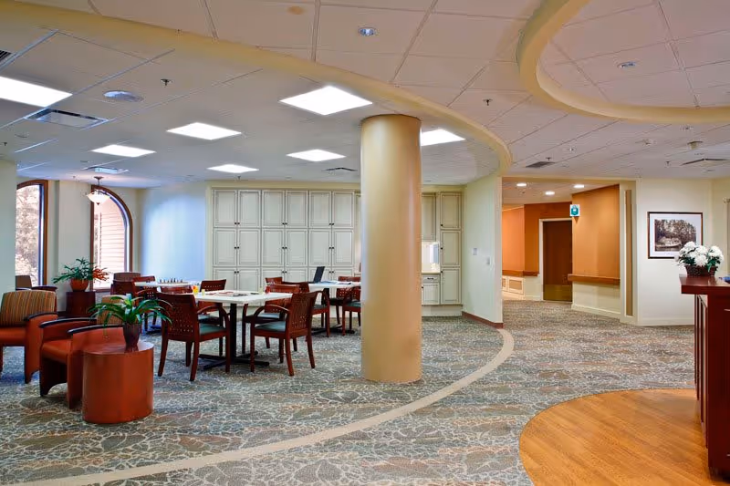 A spacious and well-lit common area in a senior living facility featuring multiple tables and chairs arranged for socializing or activities, comfortable armchairs with small side tables and potted plants, a large central column, and a patterned carpet with a curved design. The walls are light-colored with some cabinetry and a framed picture, and there is a hallway leading to other rooms.