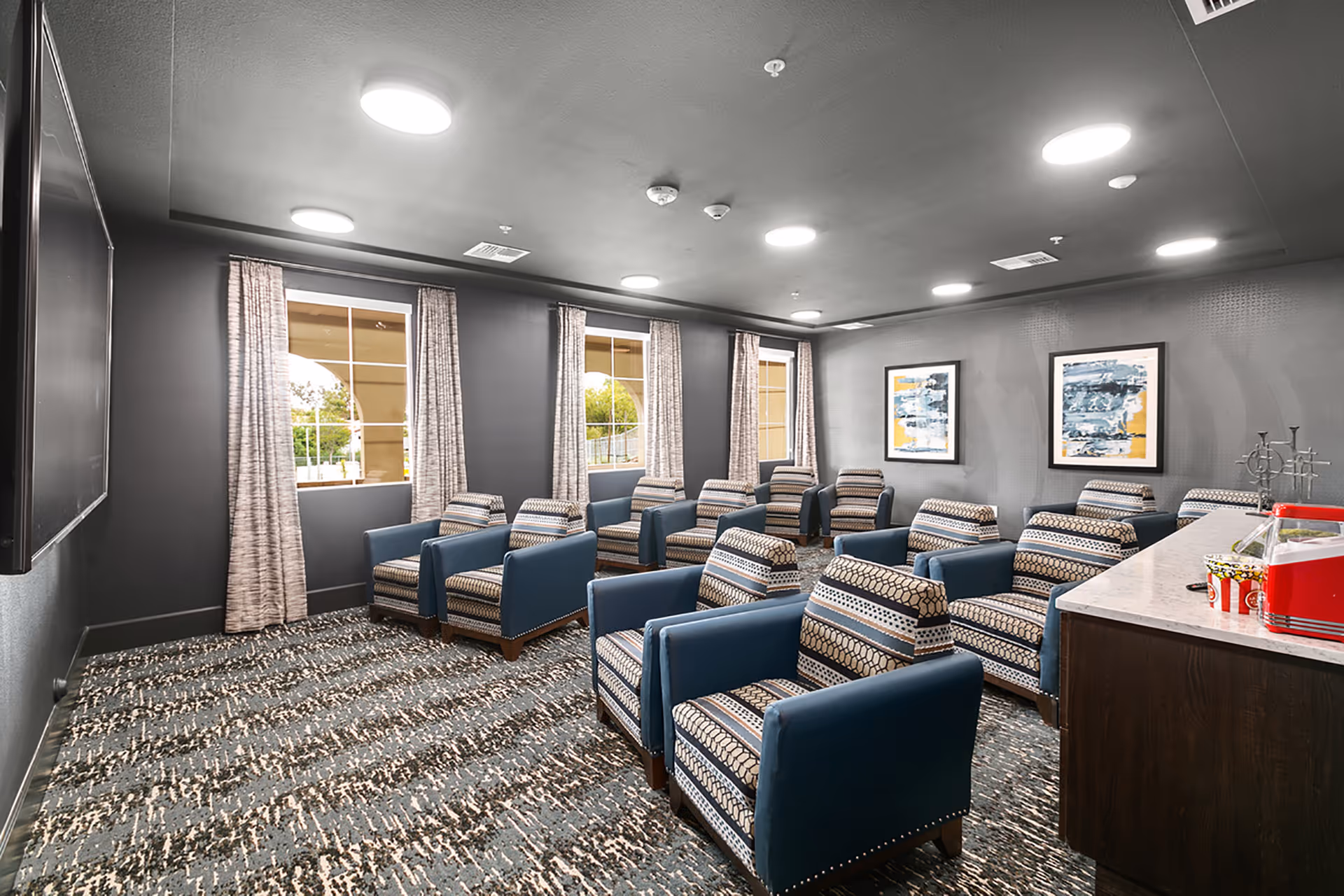 A small theater-style room with multiple rows of patterned armchairs facing a large flat screen TV mounted on a dark gray wall. The room has three windows with beige curtains, abstract artwork on the walls, and a popcorn machine on a counter to the right.