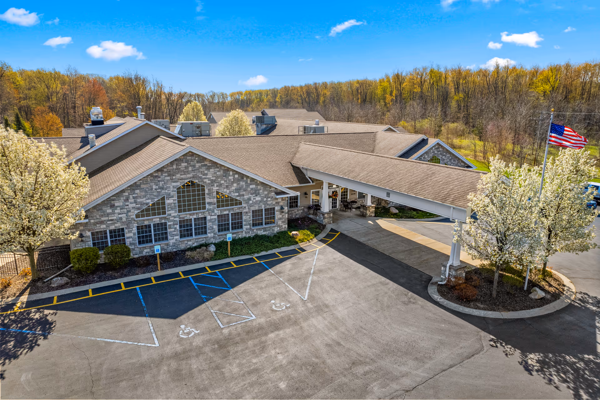 Aerial view of a stone-clad senior living building with a covered entrance, parking lot, and flowering trees.