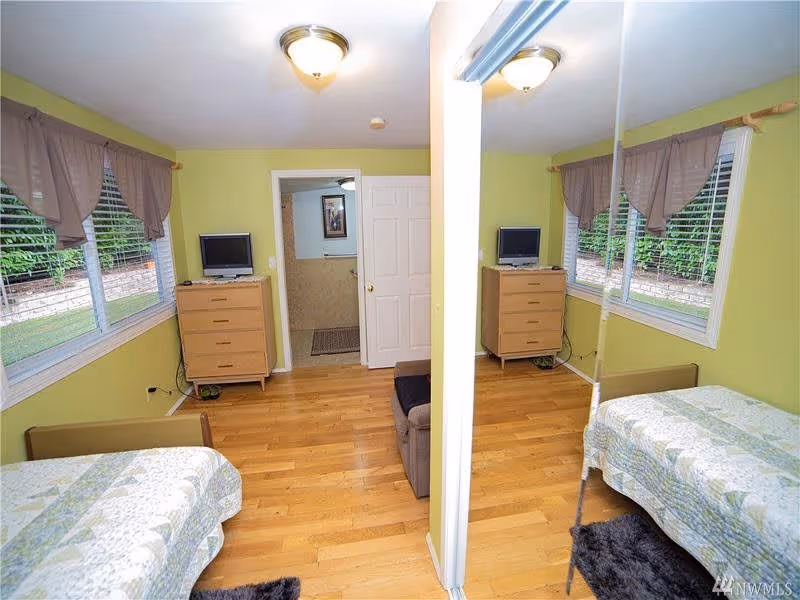 A small bedroom with light green walls and wooden floor. The room has a single bed with a patterned quilt, a wooden dresser with a small TV on top, and a window with mauve curtains. A mirrored closet door reflects the room. An open door leads to a bathroom with beige tiled walls.