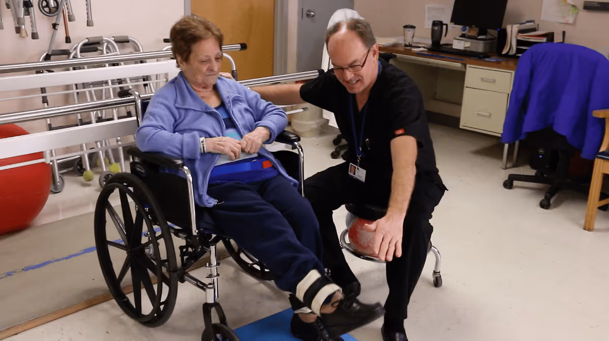 An elderly woman in a wheelchair is assisted by a healthcare professional in a rehabilitation room. The woman has a strap around her waist and an ankle weight on her leg, while the professional is seated on a stool and appears to be guiding her leg exercise. The room contains medical and rehabilitation equipment, including walkers and exercise balls.