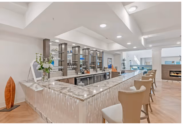Bright modern communal bar and lounge with a marble countertop, upholstered bar stools, shelving stocked with glassware and bottles, and a fireplace in the background.