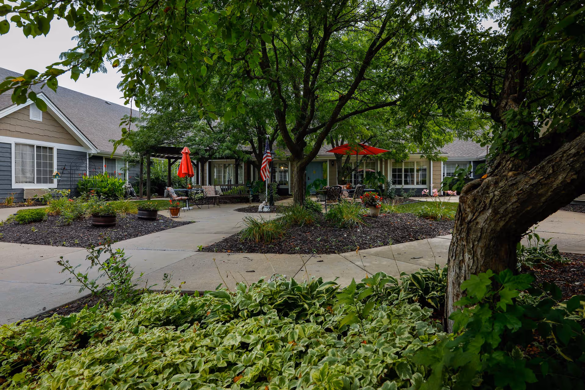 Outdoor courtyard area of Homestead Assisted Living of Leavenworth featuring a large tree in the center, surrounded by paved walkways, garden beds with plants, and seating areas with tables and red umbrellas. The building exterior is visible in the background with windows and a covered patio area.