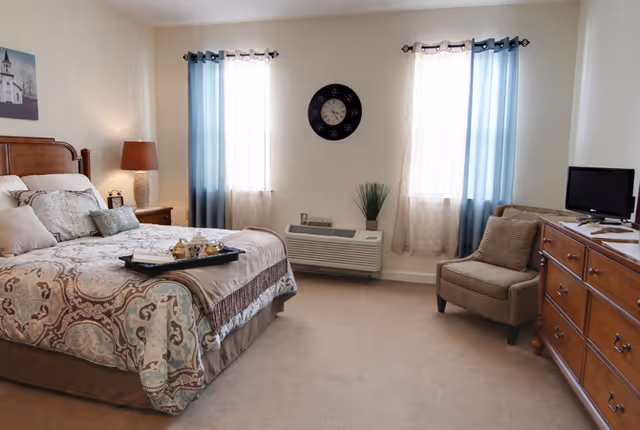 A cozy bedroom with a large bed covered in patterned bedding and multiple pillows. There is a wooden nightstand with a lamp and a small clock on the left side of the bed. Two windows with blue and white curtains are on the far wall, with a wall clock centered between them. Below the clock is a heating/cooling unit with a small plant on top. To the right, there is a cushioned armchair and a wooden dresser with a small TV on it.
