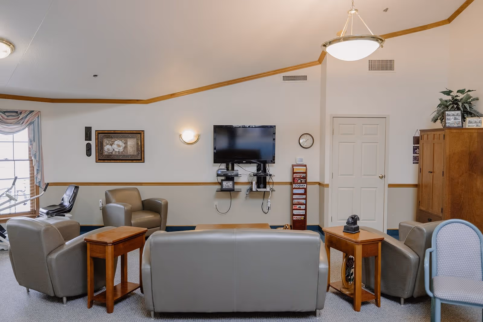 A cozy living room area in Redbud Plaza Assisted Living featuring a gray leather sofa and three matching armchairs arranged around a wooden coffee table. The room has beige walls with wood trim, a wall-mounted flat-screen TV, a clock, a wooden cabinet, and a magazine rack. There is a window with draped curtains and an exercise bike near it. The ceiling has a hanging light fixture and a wall sconce providing illumination.