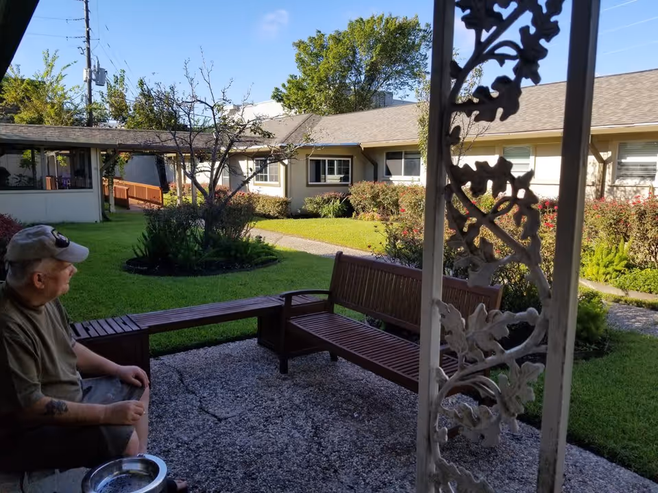 An elderly man wearing a cap and sunglasses sits on a wooden bench in a garden courtyard area of a residential facility. The courtyard has green grass, bushes, a tree, and a paved walkway. The background shows single-story buildings with windows under a clear blue sky.