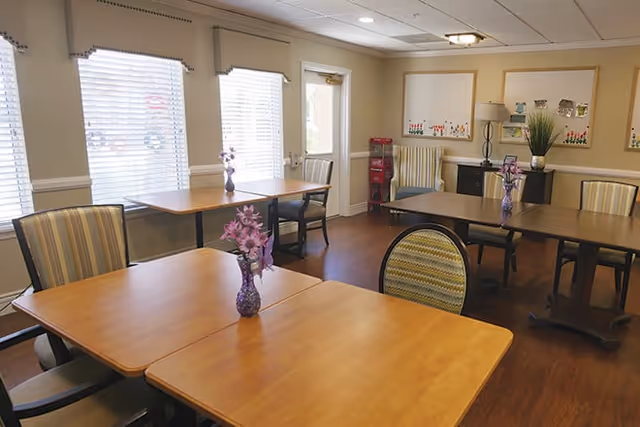 Well-lit dining room with wooden tables and chairs, small vases of purple flowers on the tables, windows with blinds, and bulletin boards on the wall.