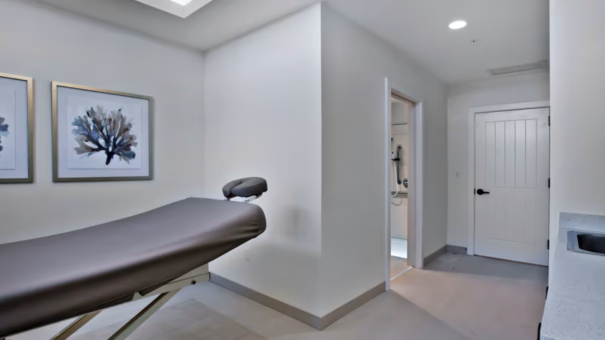 A treatment/massage table in a clean white therapy room with framed artwork and a doorway leading to a bathroom.