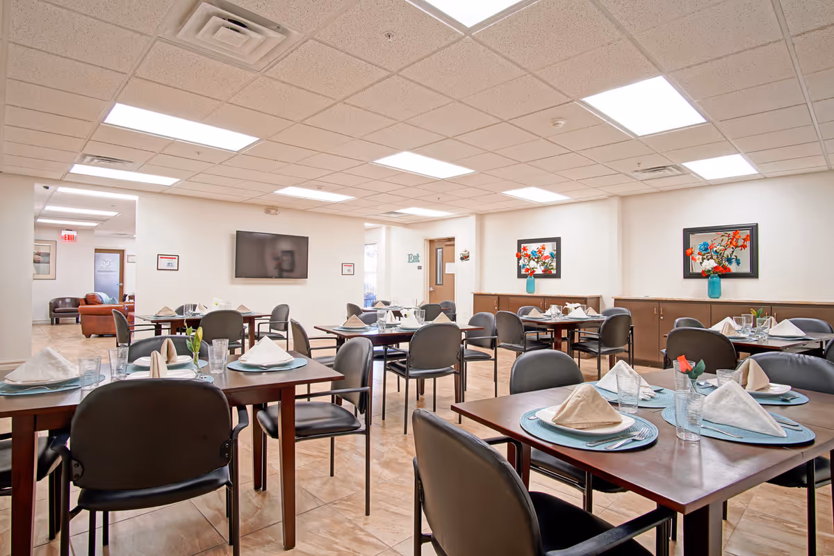 A bright dining room with multiple tables set with plates, glasses, silverware, and folded napkins. The room has a tiled floor, white walls, and a drop ceiling with fluorescent lighting. There are black chairs around the tables, colorful floral artwork on the walls, and a flat-screen TV mounted on one wall. A door and an exit sign are visible in the background.