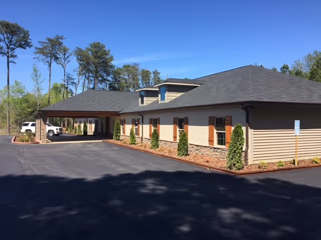 Exterior view of a single-story assisted living facility building with beige siding, stone accents, and a dark shingled roof. The building has several windows with wooden shutters and small shrubs planted along the foundation. A covered entrance is visible with a white vehicle parked nearby. The sky is clear and blue, and tall trees are in the background.