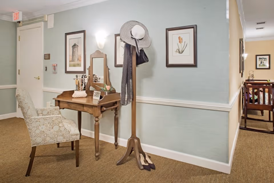 A cozy interior corner of a senior living facility with a wooden vanity table, a floral upholstered chair, and a wooden coat rack holding a striped hat, scarf, and a handbag. The walls are decorated with framed botanical and architectural prints, and soft lighting creates a warm atmosphere.