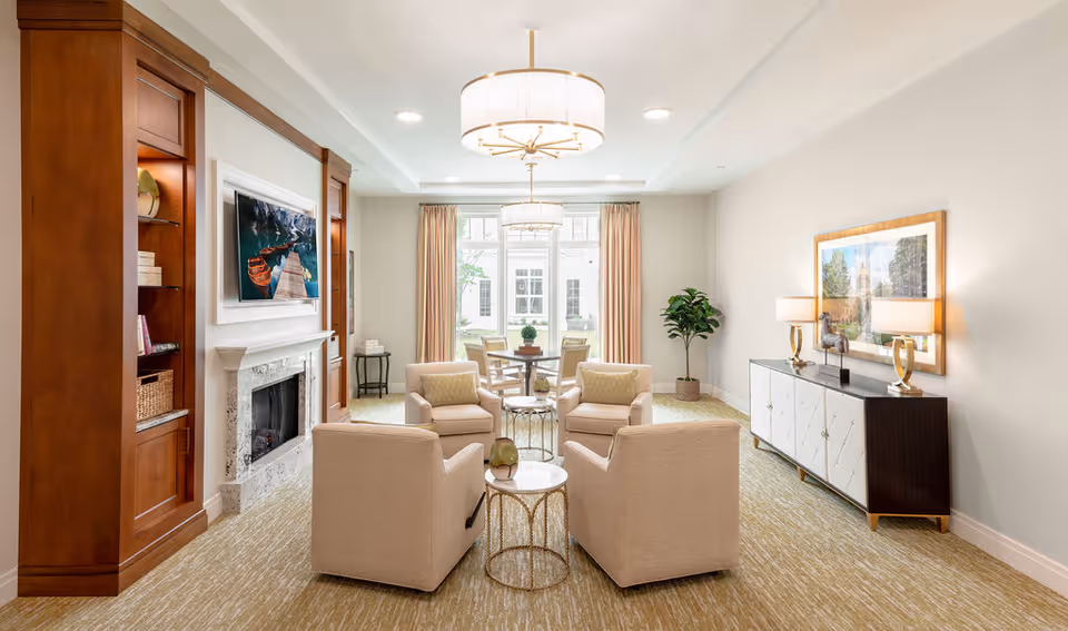A bright and elegant living room with four beige armchairs arranged around two small round tables. The room features a fireplace with a wooden built-in shelving unit on the left, a large window with peach curtains at the back, and a sideboard with two lamps and a framed picture on the right. The carpet is light-colored, and there are two modern ceiling lights.