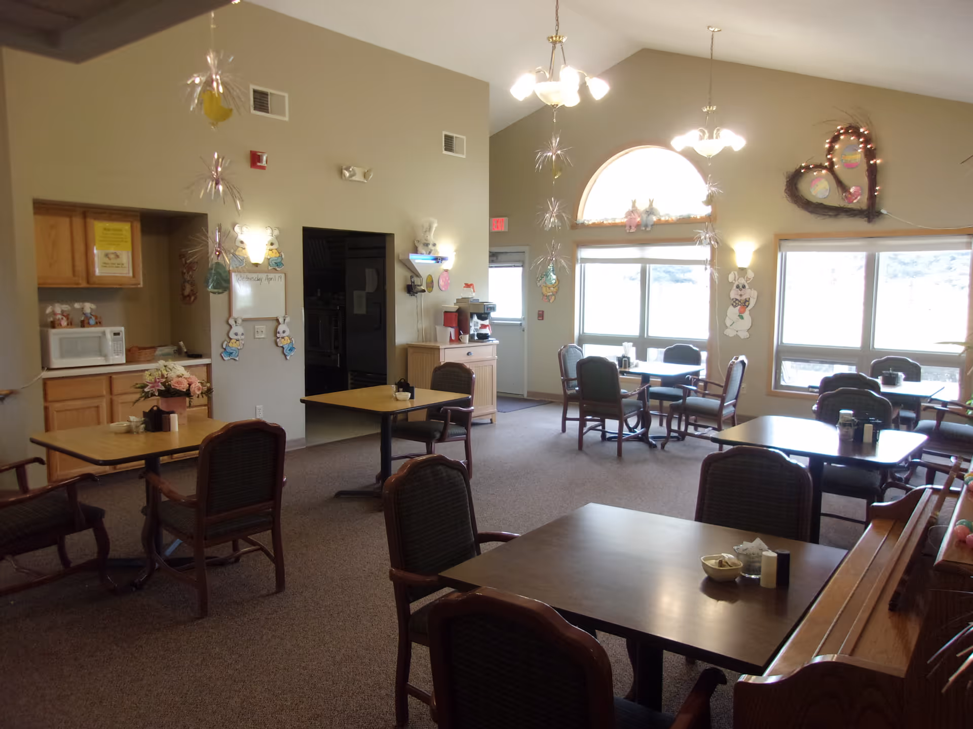 A dining area in an assisted living facility with several tables and chairs arranged around the room. There are large windows letting in natural light, wall decorations including Easter-themed bunnies and hearts, and a small kitchenette area with a microwave and coffee station. The room has a high ceiling with hanging light fixtures and festive hanging decorations.