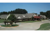Single-story residential care building with a covered entrance, circular driveway, and landscaped lawns under a clear blue sky.