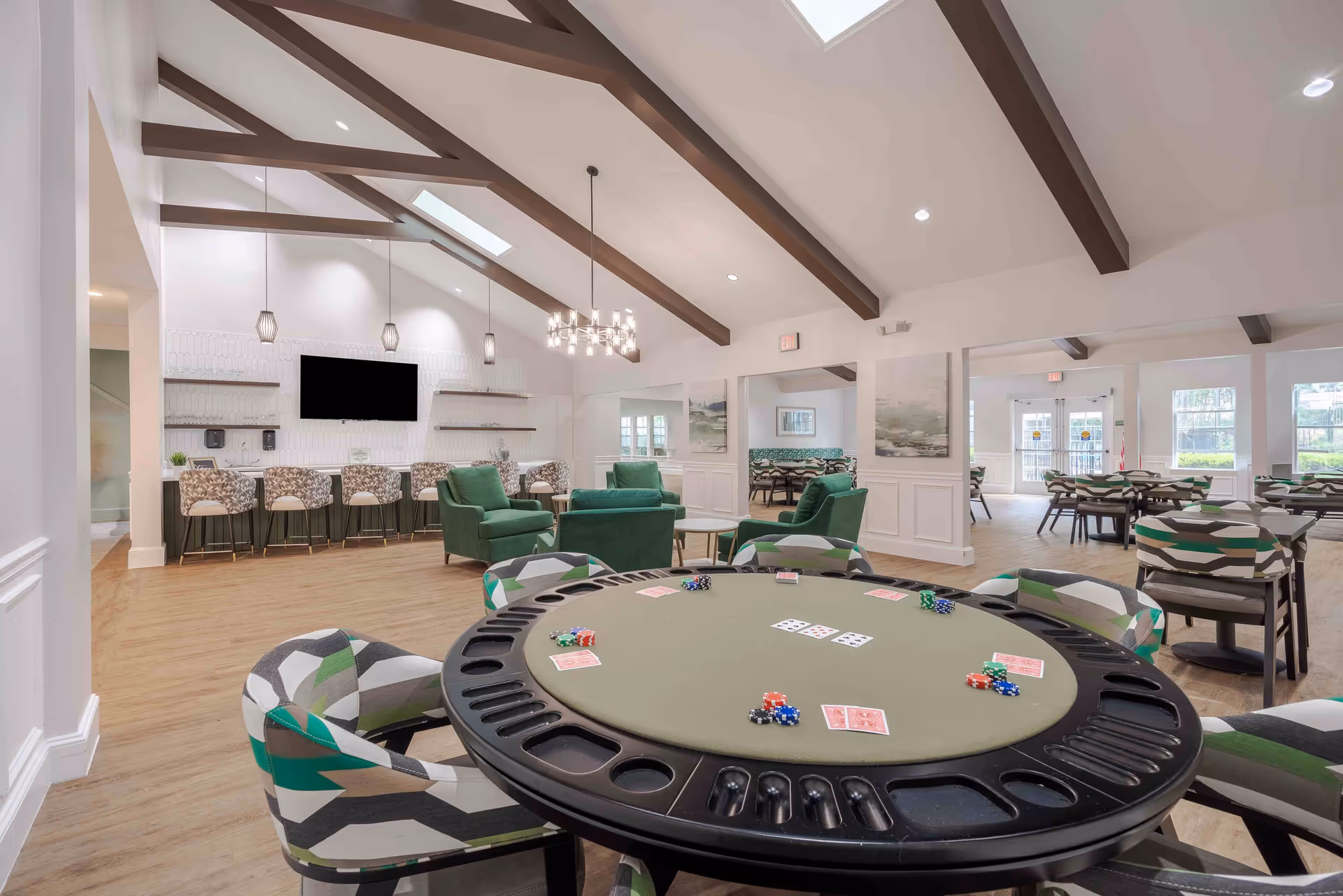 A spacious and well-lit common area with a poker table in the foreground featuring playing cards and poker chips. The room has patterned chairs around the table, green armchairs, and a bar area with high stools and a large TV mounted on the wall. The ceiling has exposed wooden beams and modern pendant lights, with additional seating and tables visible in the background near large windows and glass doors.