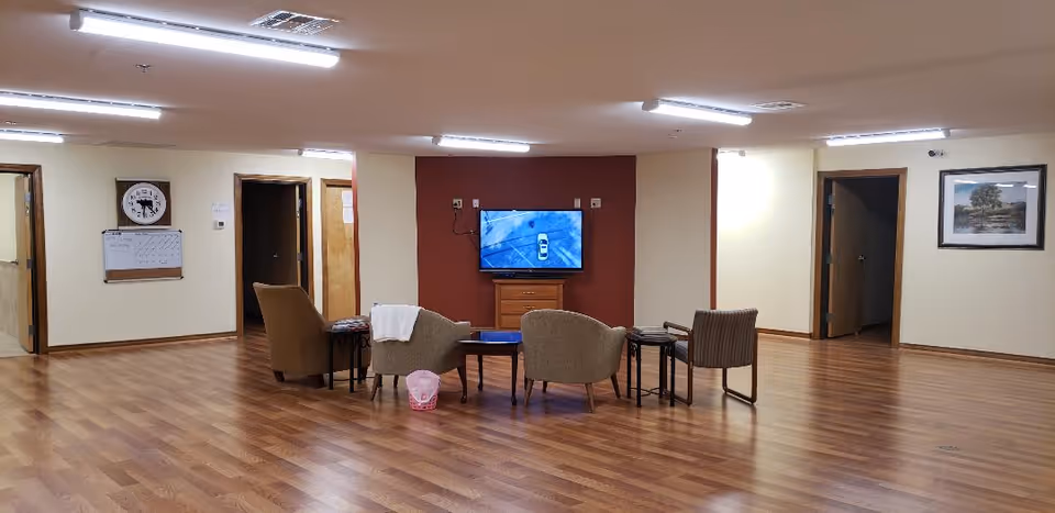 A spacious room with wooden flooring and beige walls featuring a small seating area with four chairs arranged around a coffee table facing a wall-mounted TV. The TV is mounted on a red accent wall with a wooden cabinet below it. There are multiple doorways leading to other rooms, a clock and whiteboard on one wall, and a framed picture on another wall. The room is well-lit with ceiling lights.
