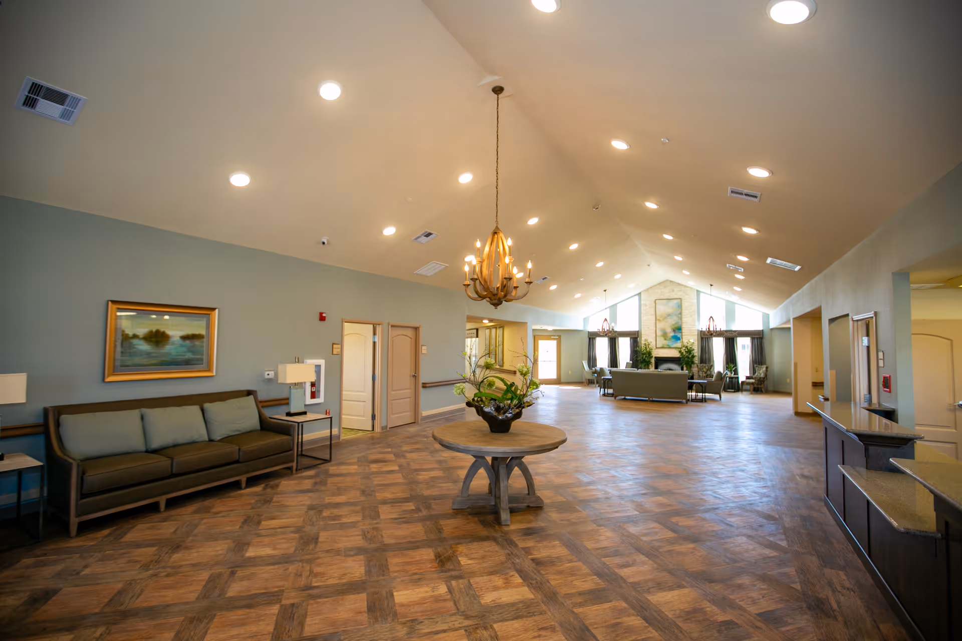 Spacious, well-lit senior living facility common area with a vaulted ceiling, chandelier, central round table with a plant, and seating along the walls and in the distance.