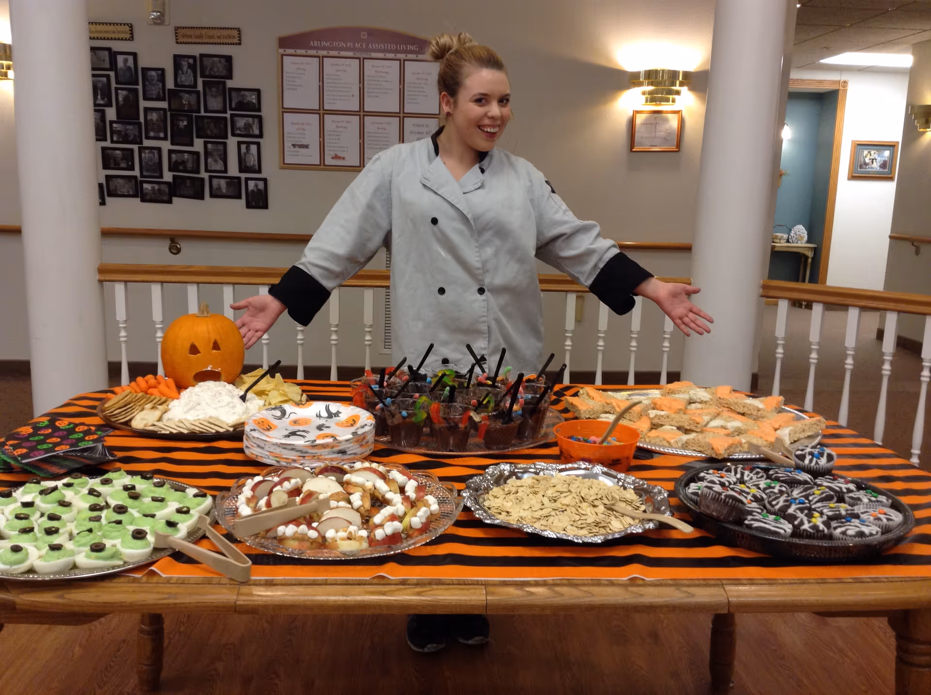 A woman in a chef's coat stands behind a table covered with a black and orange striped tablecloth, displaying a variety of Halloween-themed snacks and treats including a carved pumpkin, crackers with dip, green frosted cookies with olives, apple slices with marshmallows, cupcakes decorated with Halloween designs, and other assorted snacks. The setting appears to be an indoor common area with framed photos and notices on the wall behind her.