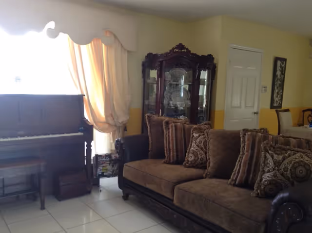 Living room with a brown upholstered sofa, ornate glass display cabinet, upright piano, and a curtained window.