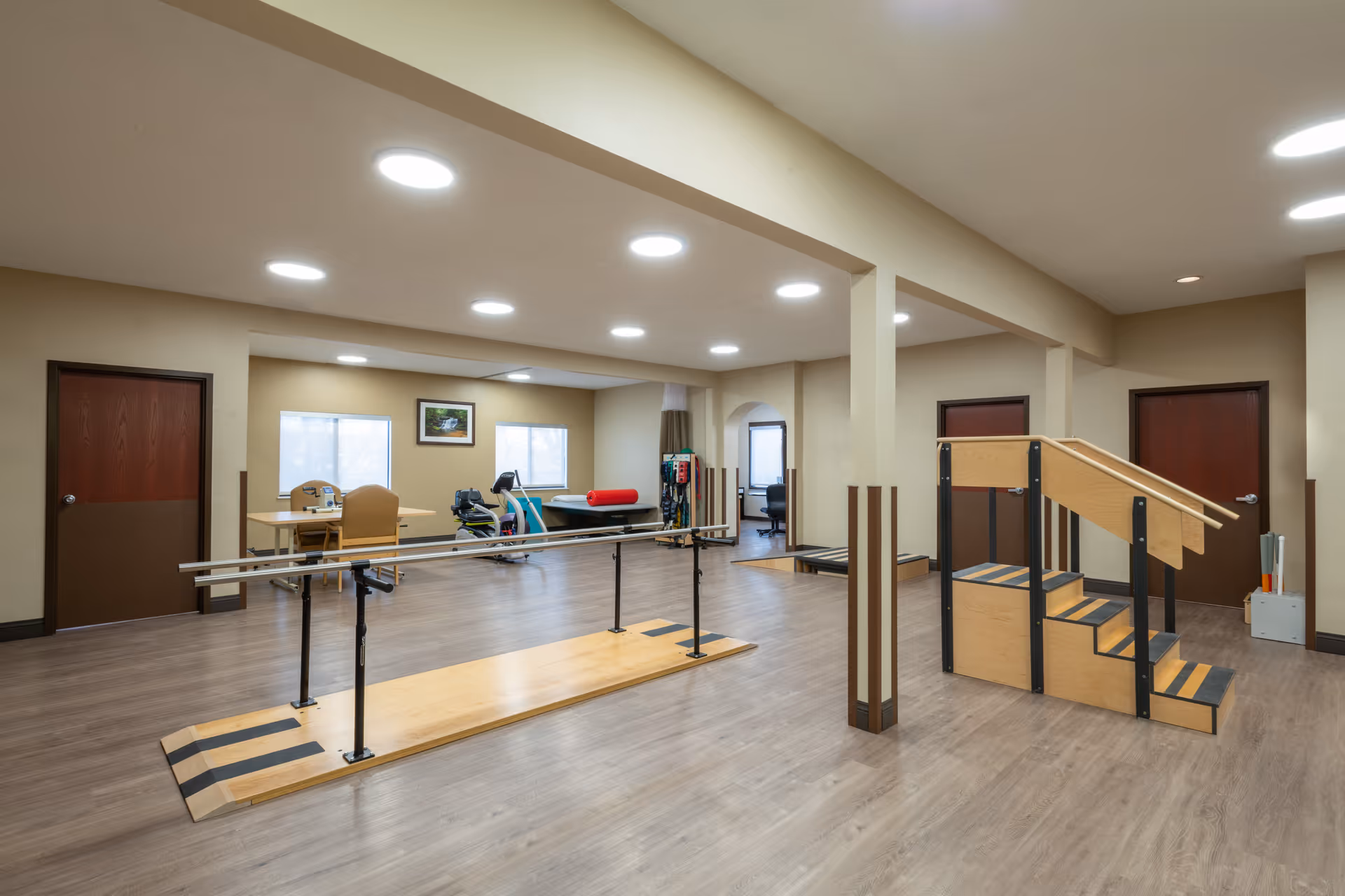 A spacious rehabilitation room with wooden flooring, featuring parallel bars for walking exercises and a wooden staircase with handrails for physical therapy. The room has beige walls, multiple ceiling lights, two windows, and several doors. In the background, there is exercise equipment and a table with chairs.