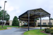 Entrance of a senior living facility with a covered drop-off area supported by metal beams, surrounded by landscaped greenery and trees under a cloudy sky.