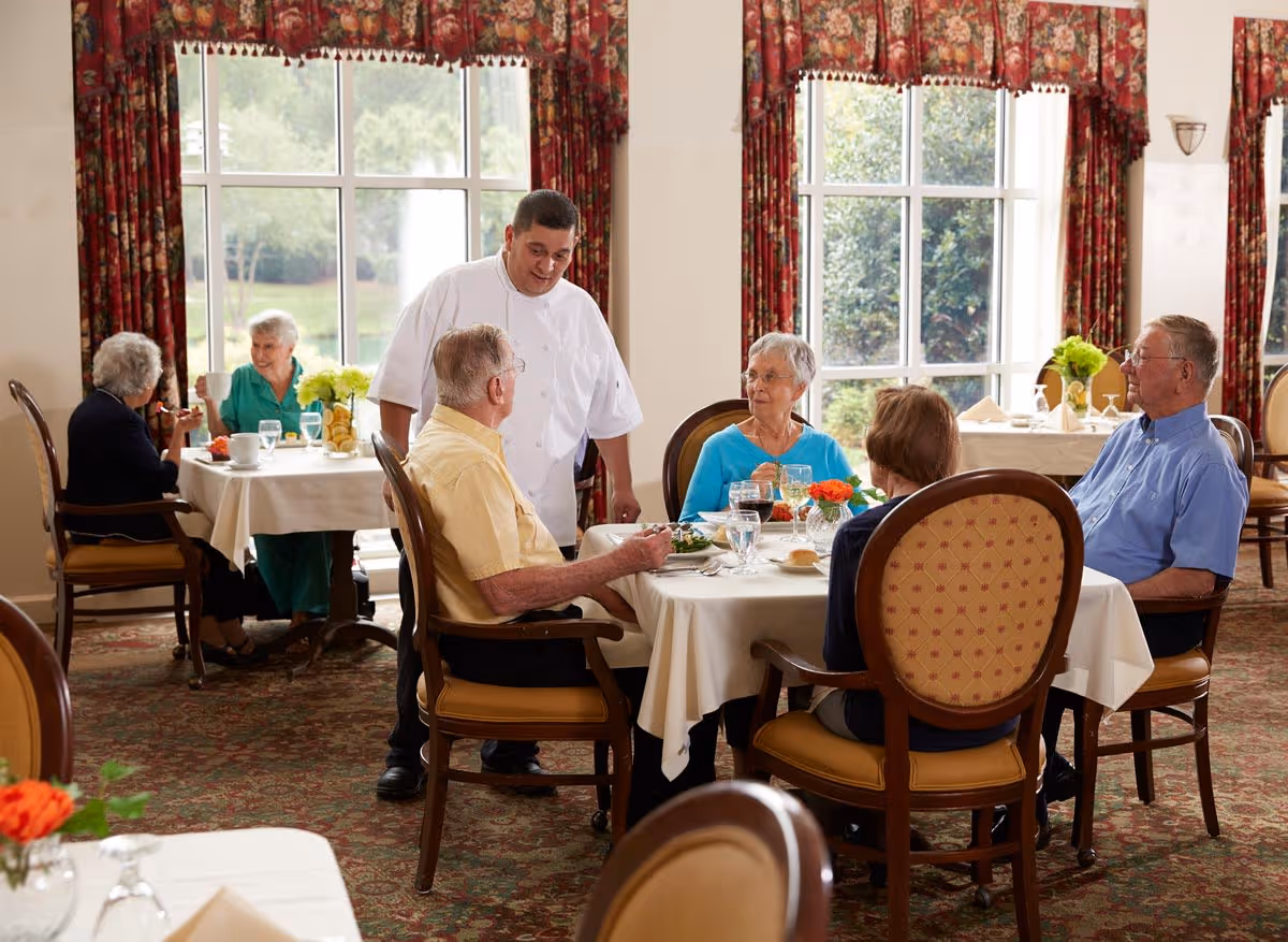 A dining room in a senior living facility with elderly residents seated at tables covered with white tablecloths. A staff member in a white uniform is interacting with a group of four seniors at one table. Large windows with floral curtains let in natural light, and the room has a warm, inviting atmosphere.