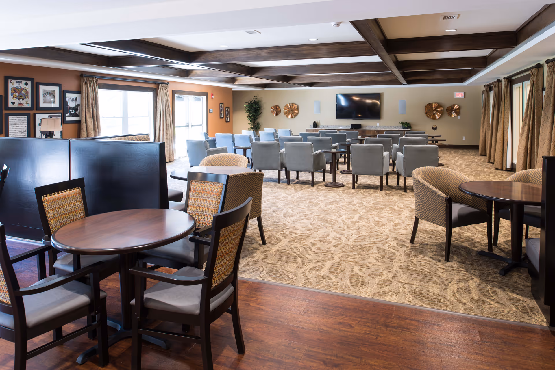 A spacious common area with multiple seating arrangements including round tables with chairs and rows of armchairs facing a wall-mounted flat screen TV. The room features wood beam ceiling accents, patterned carpet, framed artwork on the walls, large windows with curtains, and warm lighting.