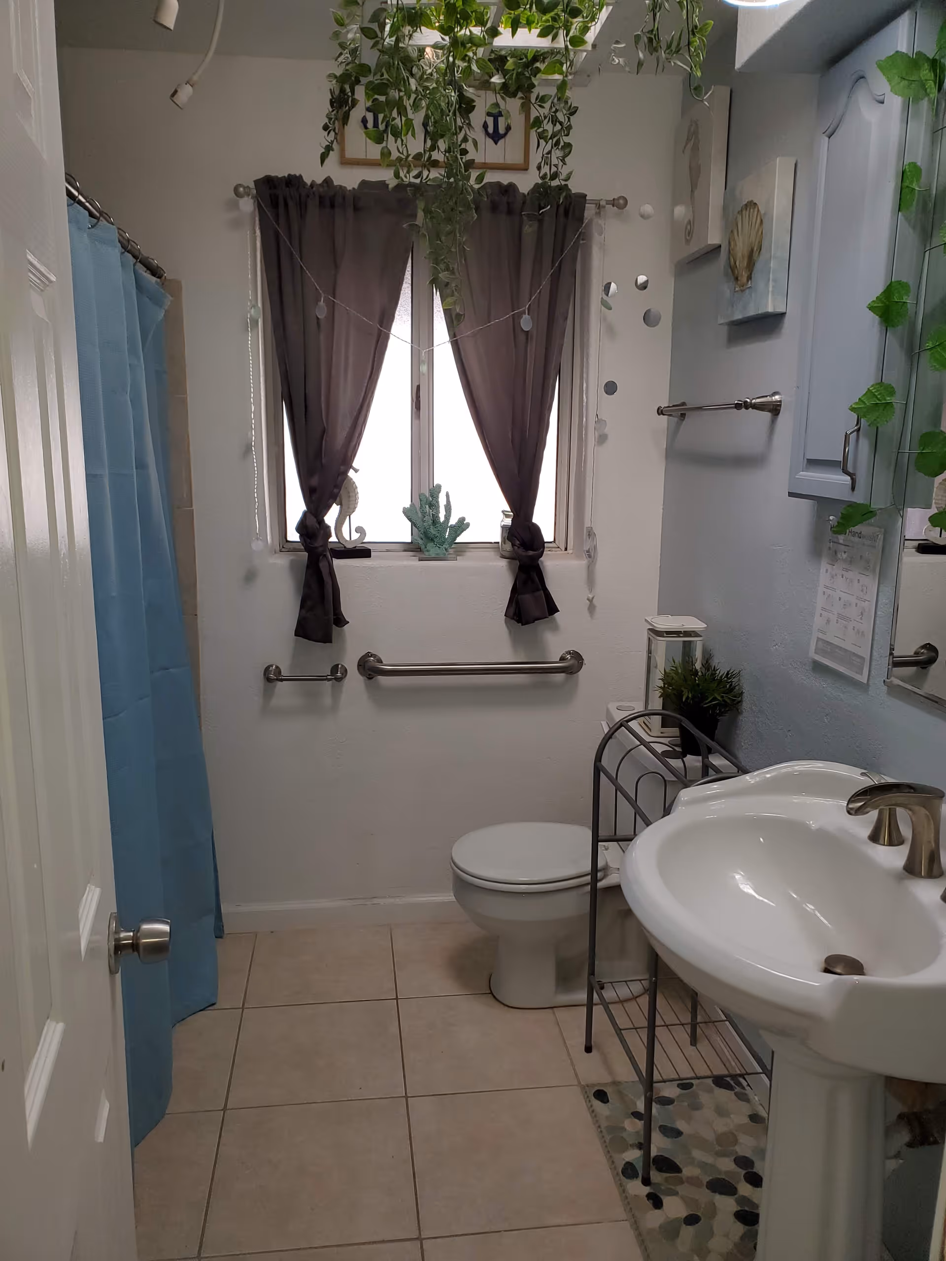 A small bathroom with a white pedestal sink, a toilet, and a shower with a blue curtain. There are two dark curtains on the window above the toilet, with hanging green plants and decorative items on the windowsill. The walls are light-colored, and there are grab bars installed near the toilet. A small metal rack holds a plant and other items next to the sink.