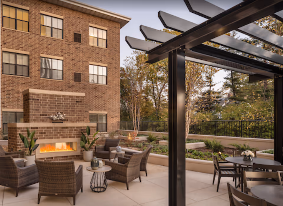 Outdoor patio area with a brick fireplace, wicker chairs, small tables with decorative items, a pergola structure, and surrounding greenery and trees.