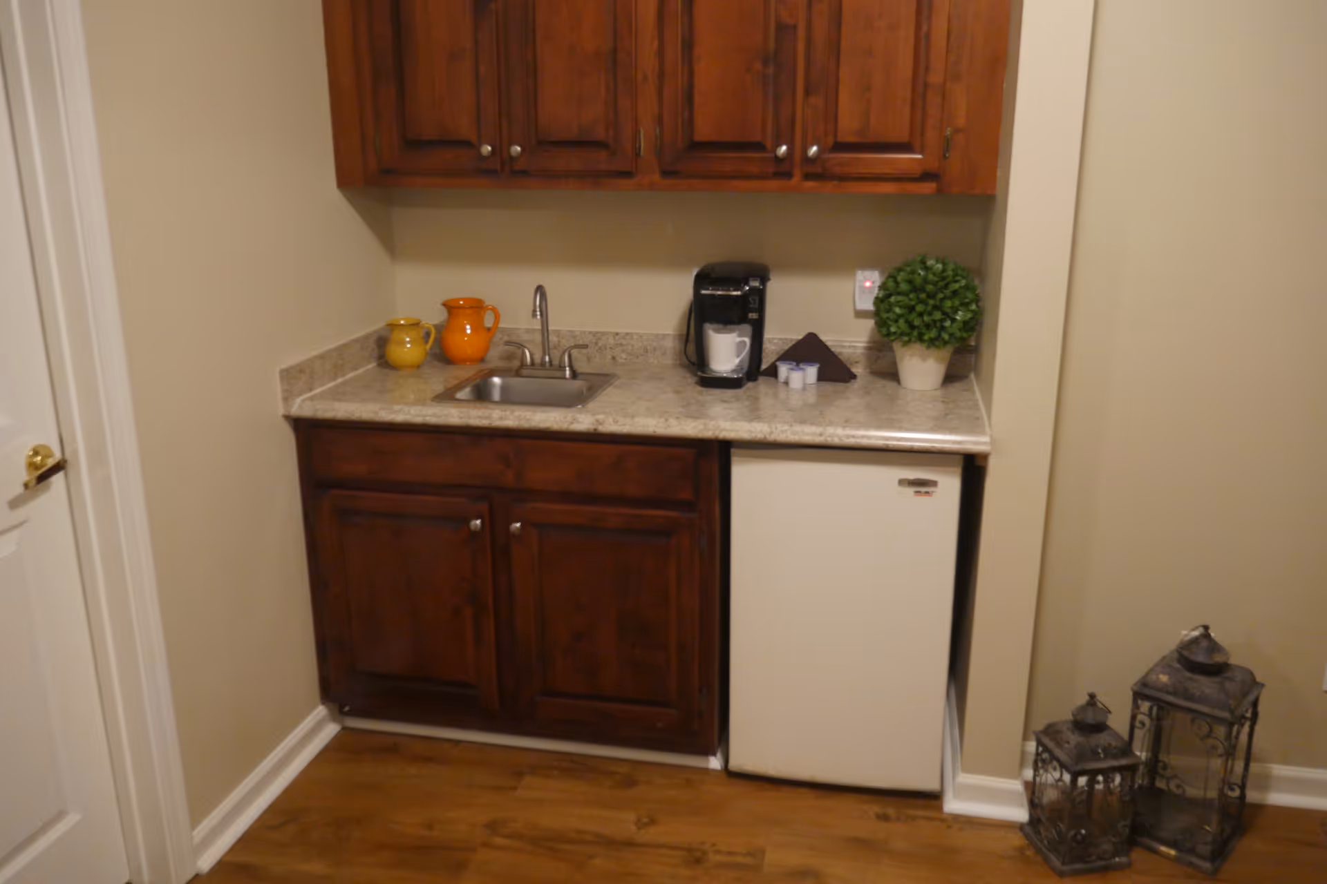 A small kitchenette with wooden cabinets, a countertop with a sink, a coffee maker with a mug, two small pitchers, a potted plant, and a mini refrigerator. Two decorative lanterns are placed on the floor to the right.