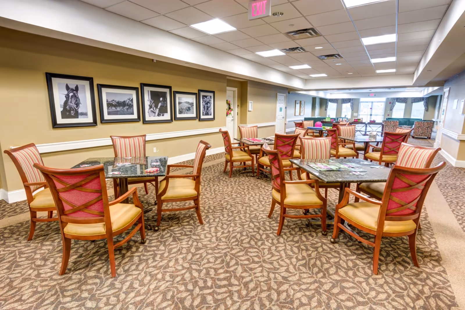 Common dining/activity room with multiple tables and upholstered chairs, patterned carpet, framed black-and-white photos on the wall, and a seating area in the background.