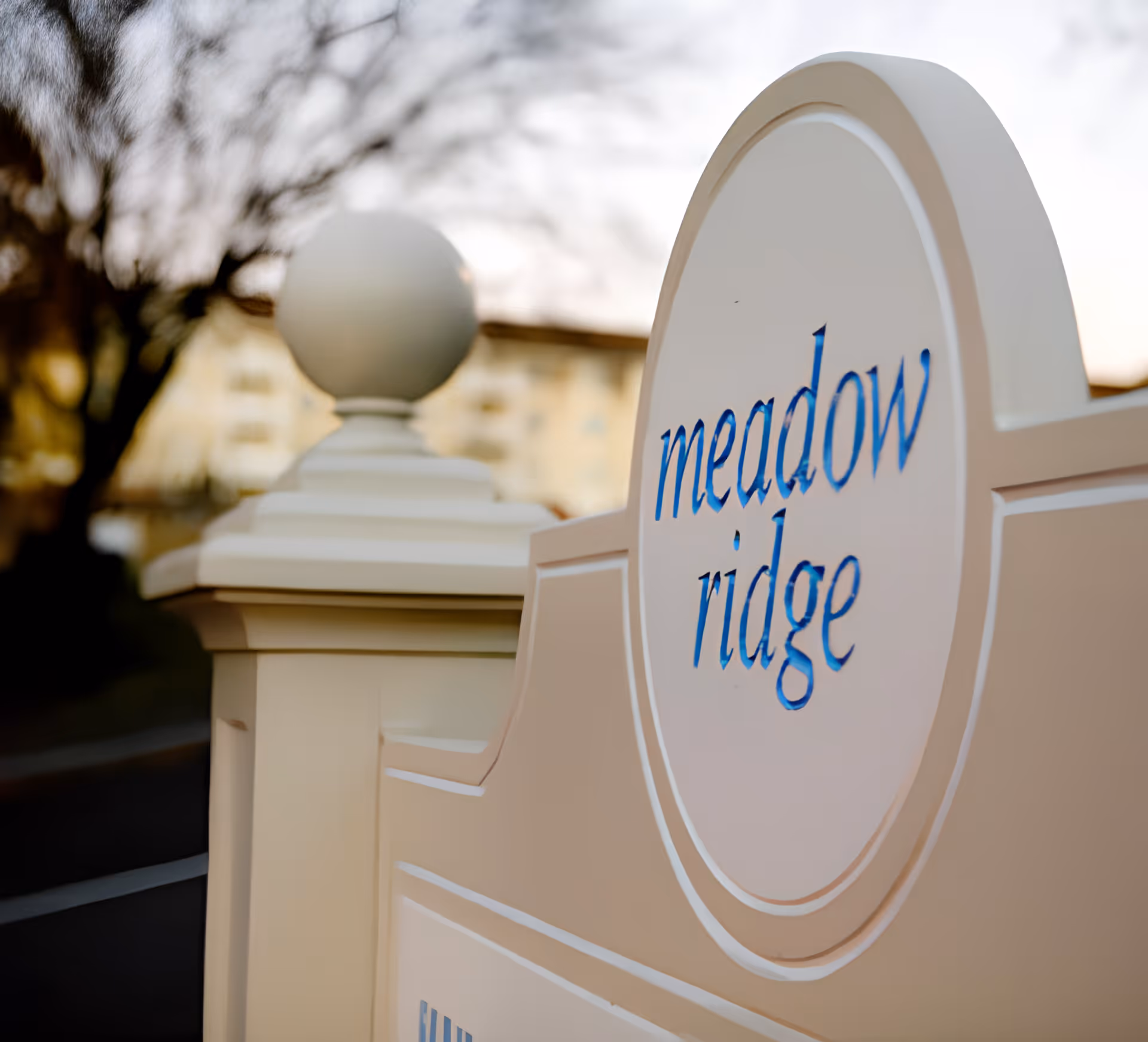 Close-up of a white outdoor sign with blue lettering that reads 'meadow ridge,' with a blurred background of trees and a building.