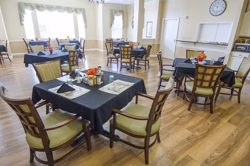 A dining room with several square tables covered with black tablecloths, each set with placemats, napkins, glasses, and small flower arrangements. The room has wooden flooring, cream-colored walls, and large windows with valance curtains allowing natural light to enter.