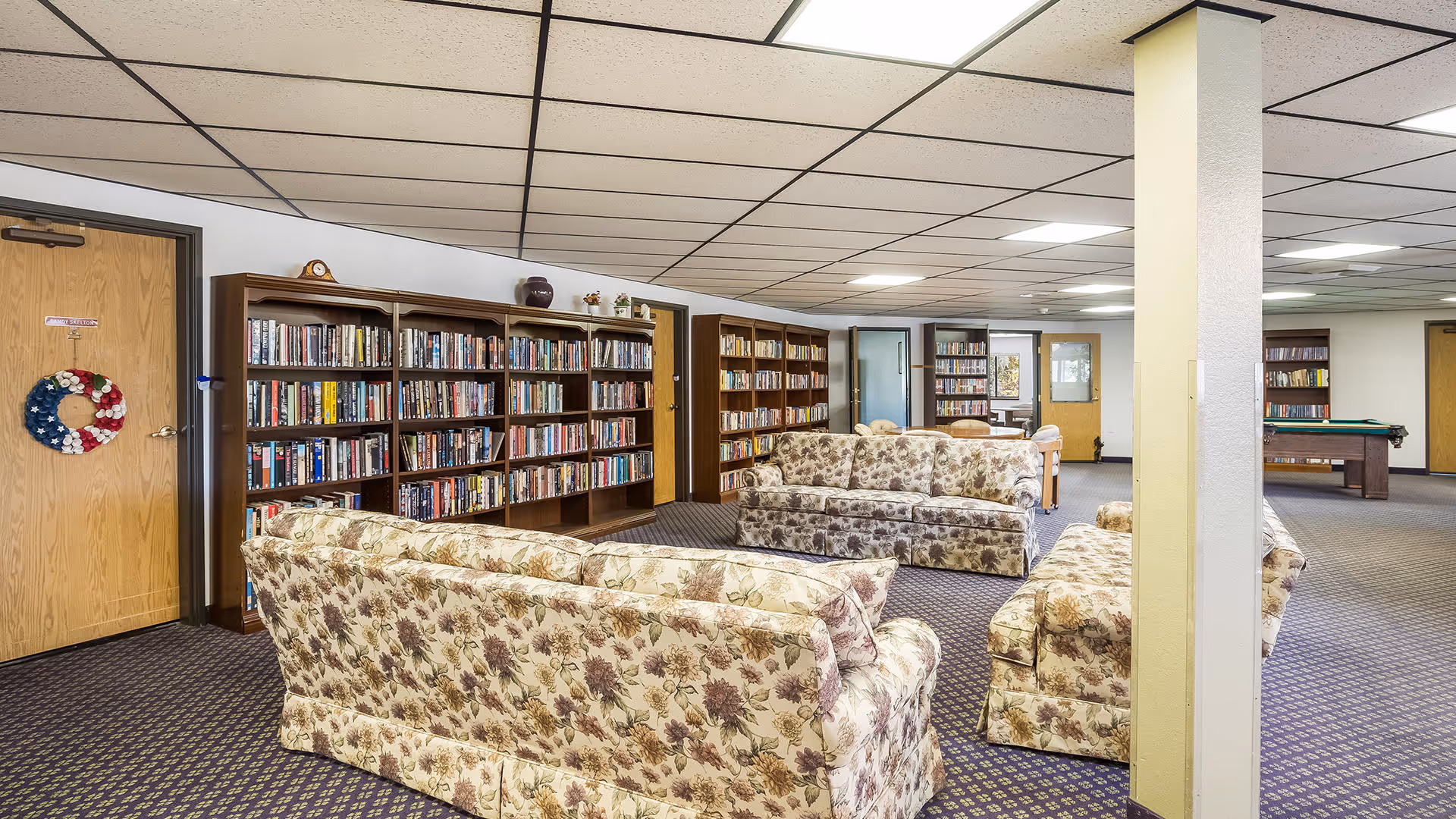 Spacious common room with floral-upholstered sofas, bookshelves lining the wall, and a pool table in the background.