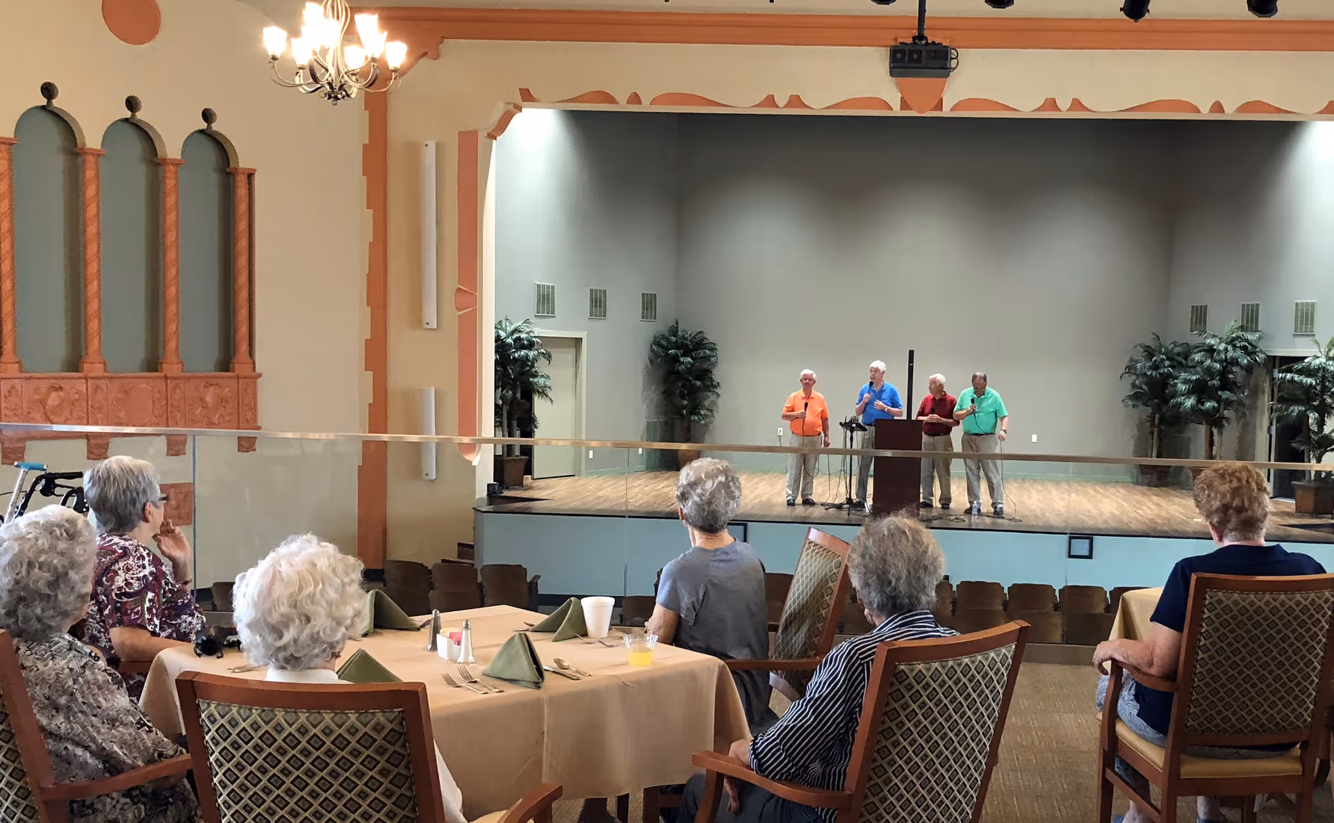A group of elderly people seated at tables with beige tablecloths and green napkins, watching four elderly men standing on a stage speaking into microphones in a large room with decorative walls and chandeliers.