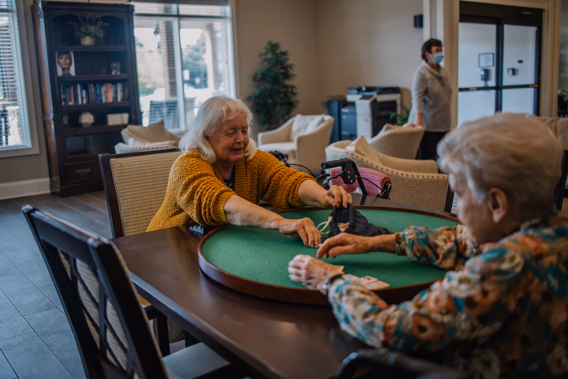 Two elderly women sitting at a round green felt table in a common area, one wearing a mustard yellow sweater and the other in a floral blouse, with a woman in a mask standing in the background near beige armchairs and a bookshelf.