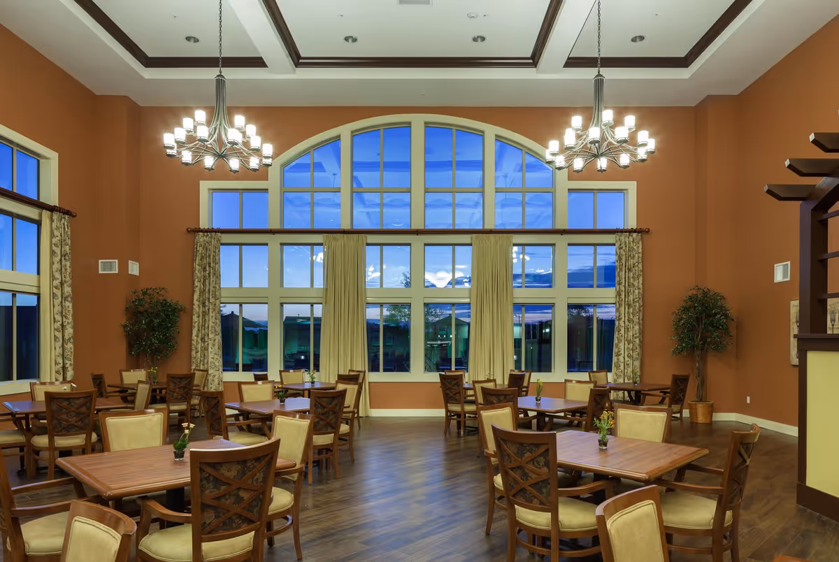 A spacious dining room with multiple wooden tables and chairs arranged neatly. The room features large arched windows with curtains, two modern chandeliers hanging from a high ceiling, and potted plants in the corners. The walls are painted a warm brown color, and the floor is wooden.