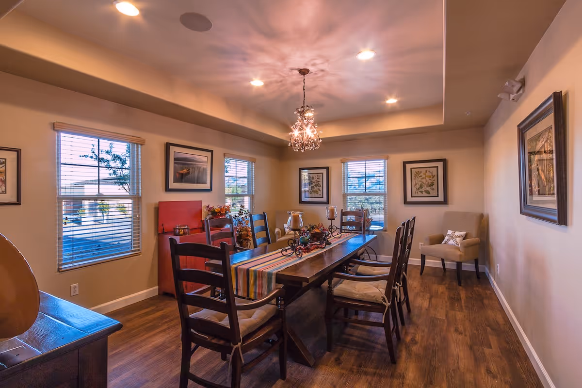 Well-lit dining room with a long wooden table and chairs, a chandelier, framed artwork, and windows with blinds.