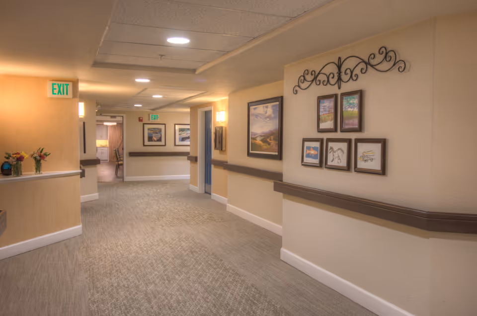 A well-lit hallway in a senior living facility with beige walls, carpeted floor, and handrails along the walls. The hallway features framed artwork and decorative wall hangings. There are exit signs visible, and a glimpse into another room with chairs and a table can be seen at the end of the hallway.