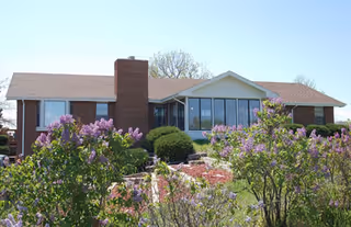 Single-story brick building with a chimney, large windows, and a sloped roof, surrounded by green bushes and blooming purple flowers under a clear sky.